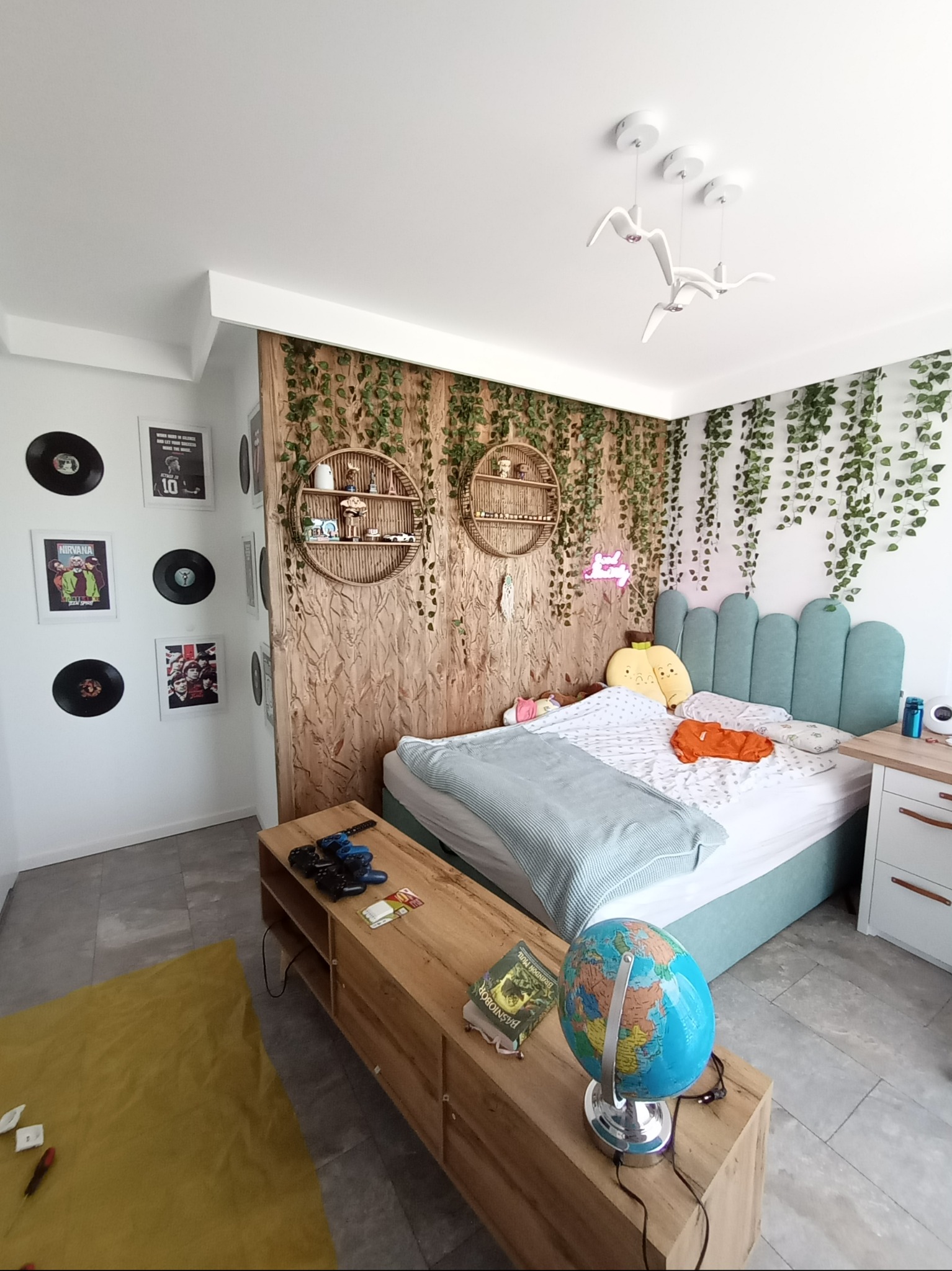 Pokój młodzieżowy z drewnianą ścianką działową udekorowaną pnączami, okrągłymi półkami i neonem, na ścianie płyty winylowe i plakaty, na szafce globus i książka.