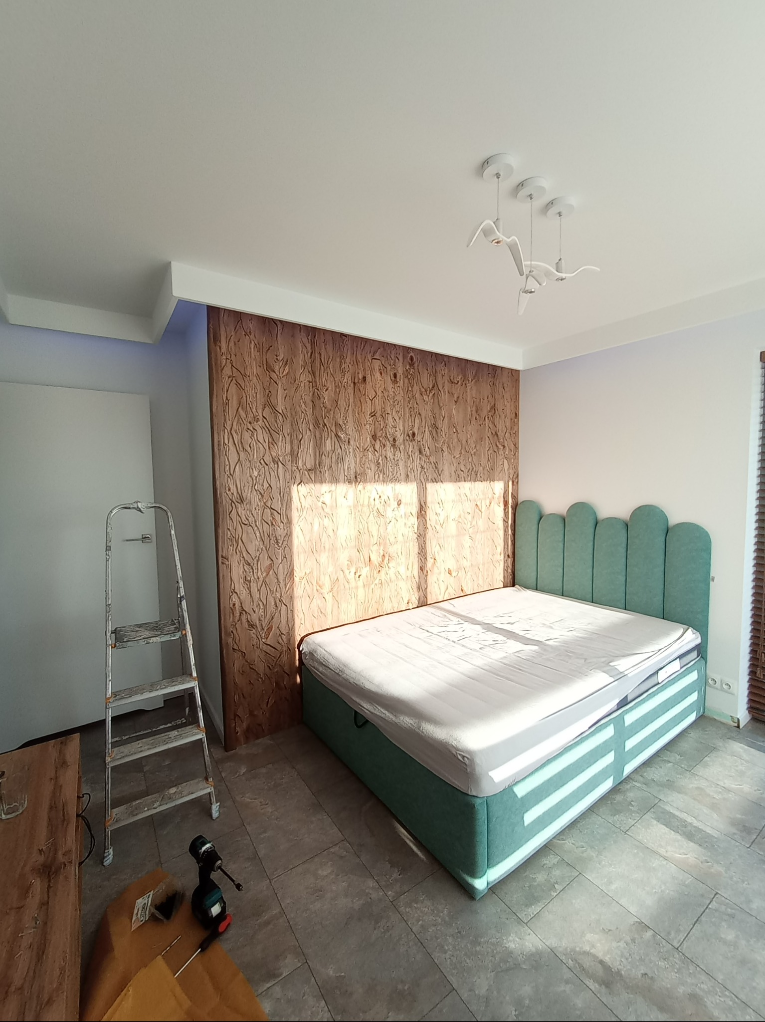 Sypialnia w trakcie wykańczania: łóżko z zielonym zagłówkiem, dekoracyjna drewniana ściana, drabina malarska i wkrętarka na podłodze, nowoczesna lampa sufitowa.