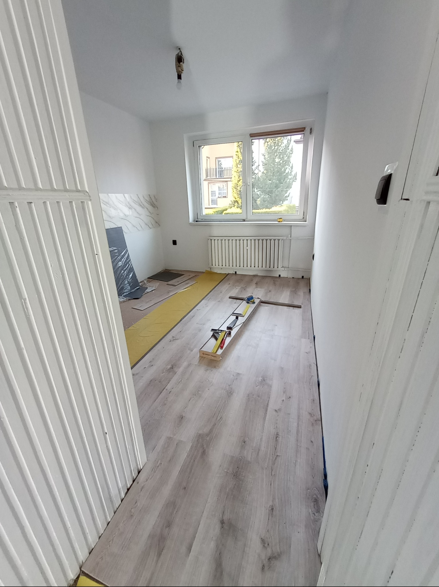 Pokój w trakcie remontu: widok na podłogę z panelami w odcieniach szarości, częściowo podłożoną folią, narzędzia do montażu paneli, białe ściany, okno z widokiem na zieleń i grzejnik.