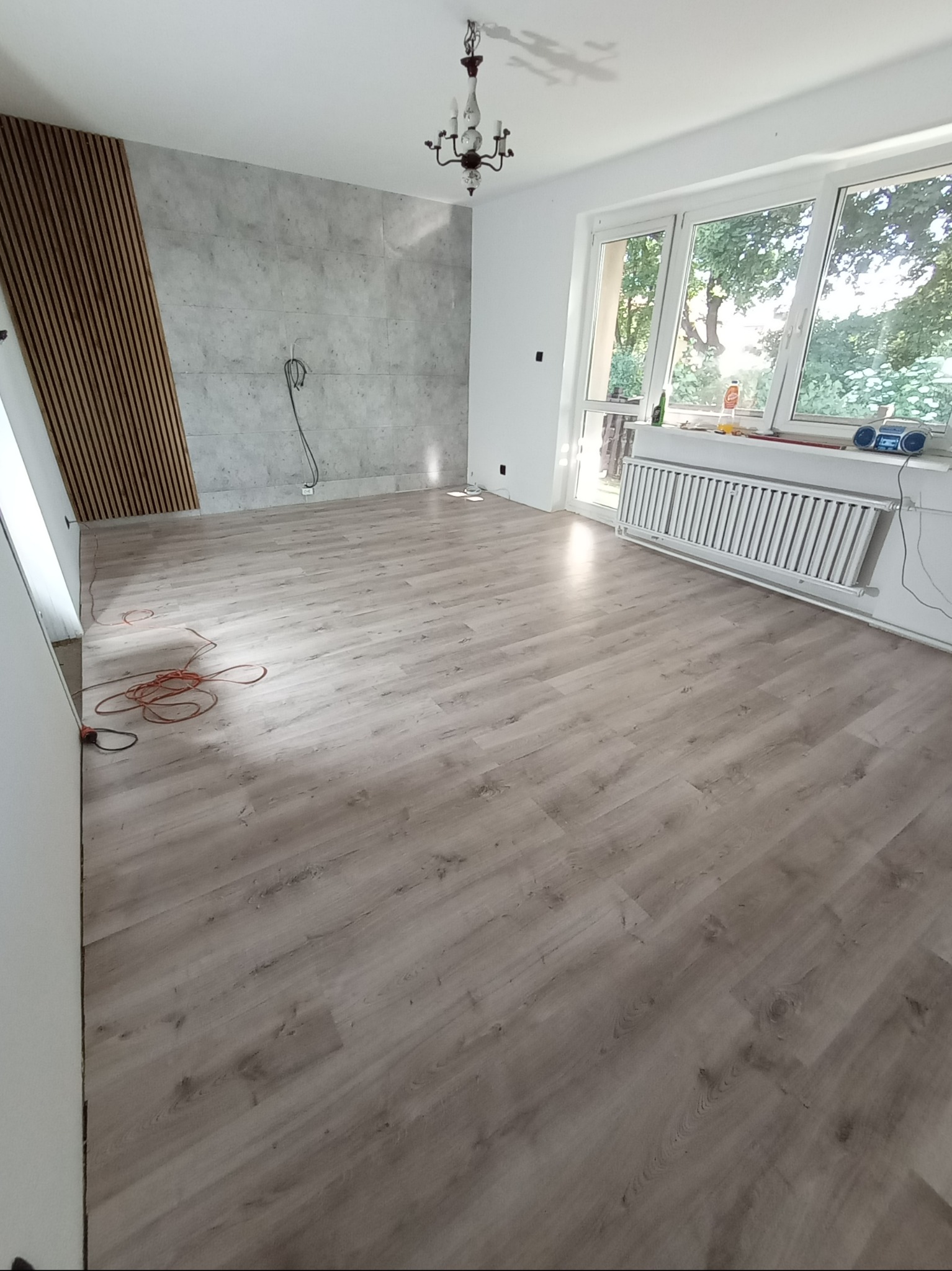 Wnętrze pokoju po remoncie z panelami ściennymi imitującymi beton i dekoracyjnymi listwami z drewna, widoczne kable wystające ze ściany, parkiet na podłodze i grzejnik pod oknem.