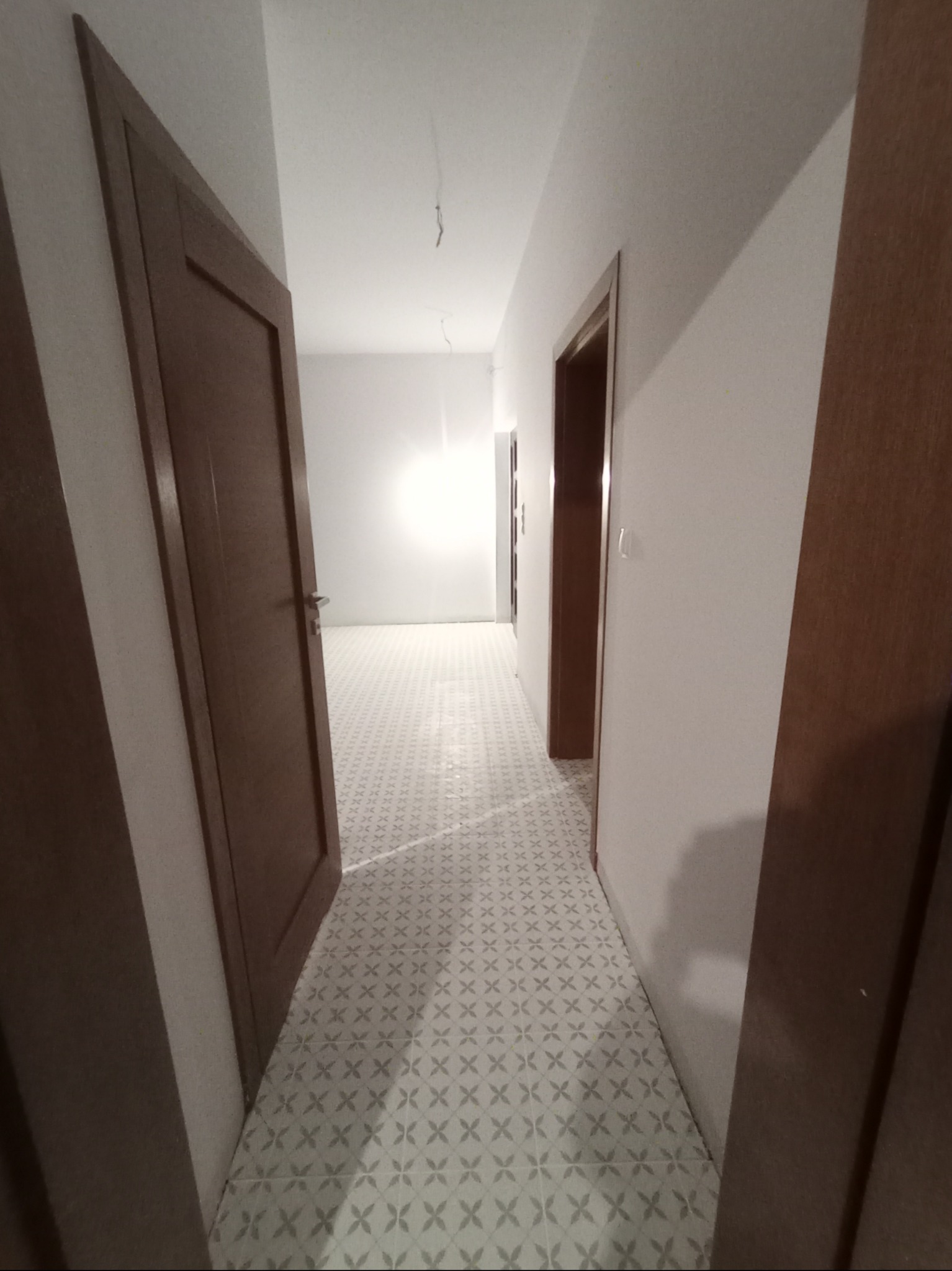 Wykończony korytarz z jasnymi ścianami, wzorzystą podłogą i drewnianymi drzwiami, widok z perspektywy wnętrza, kabel zwisający z sufitu.