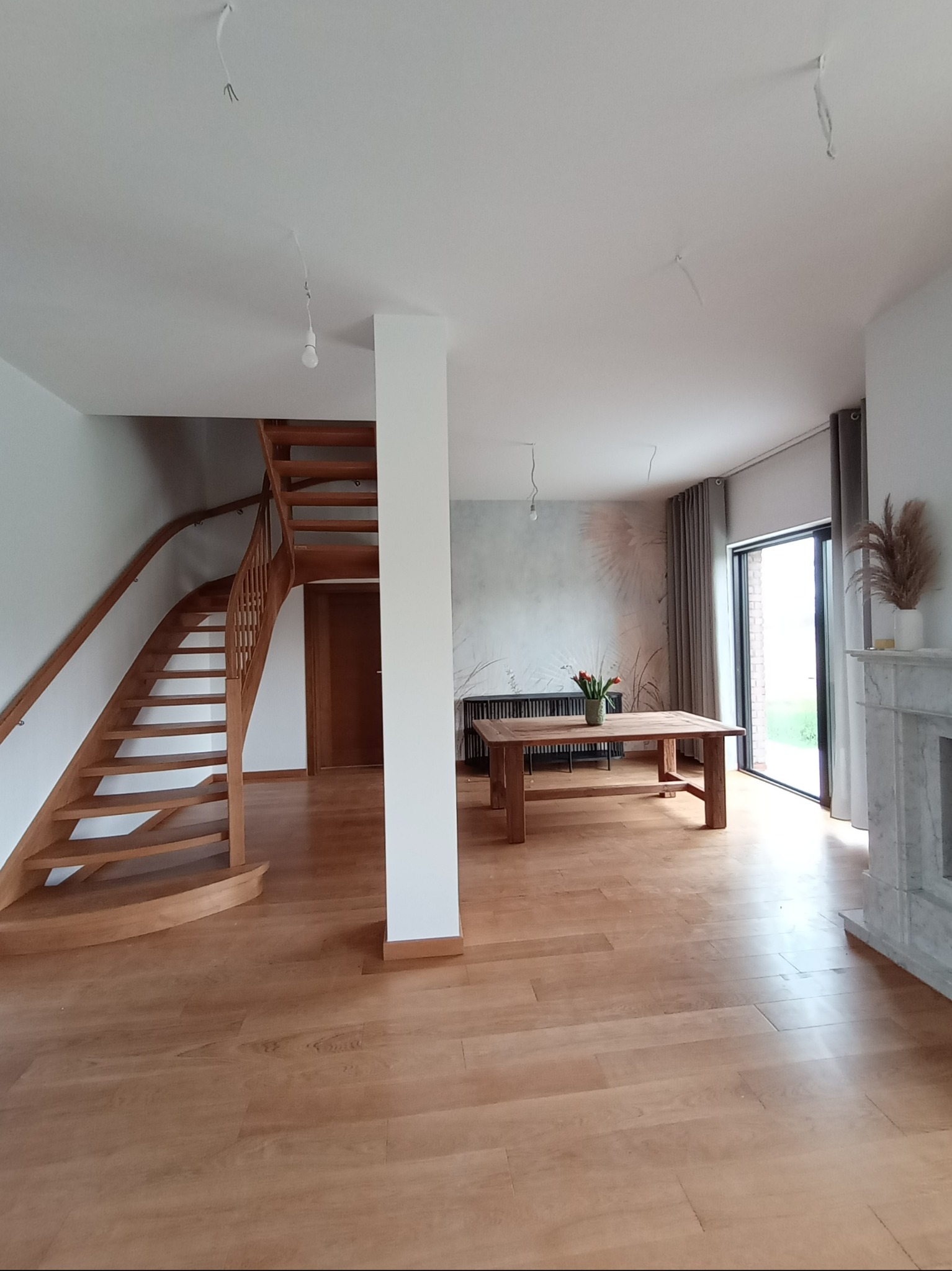 Jasne, przestronne wnętrze domu z drewnianymi schodami, parkietem i stołem. Widoczne niedokończone punkty oświetleniowe na suficie.