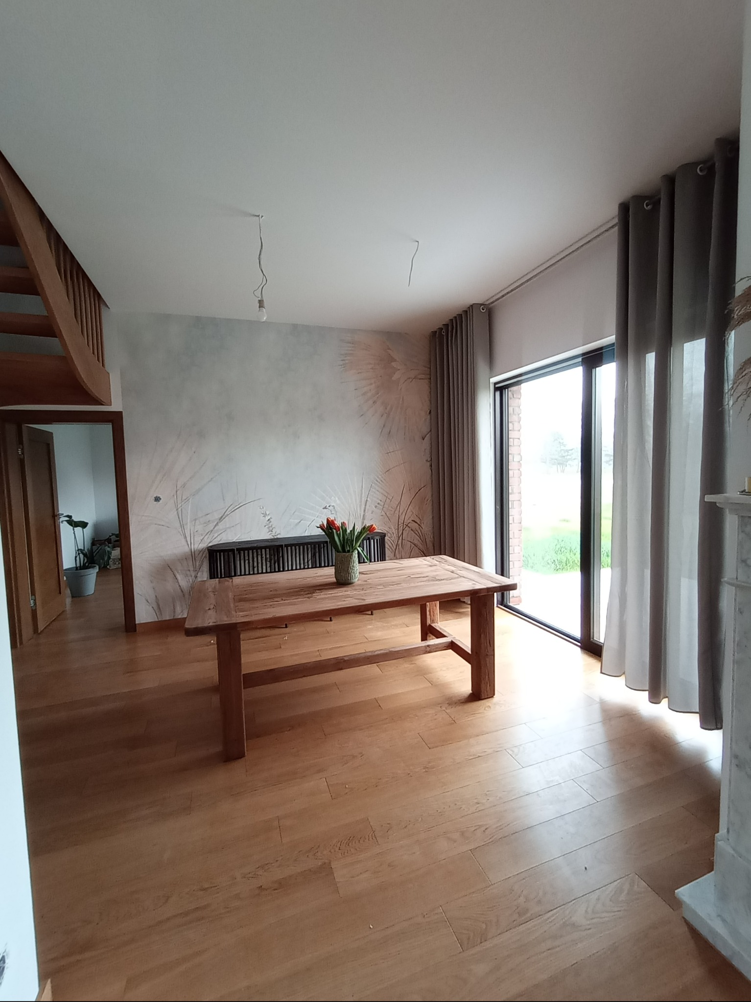 Jasne wnętrze domu z drewnianą podłogą, stołem z bukietem tulipanów, ścianą z motywem roślinnym i szarymi zasłonami przy dużym oknie.