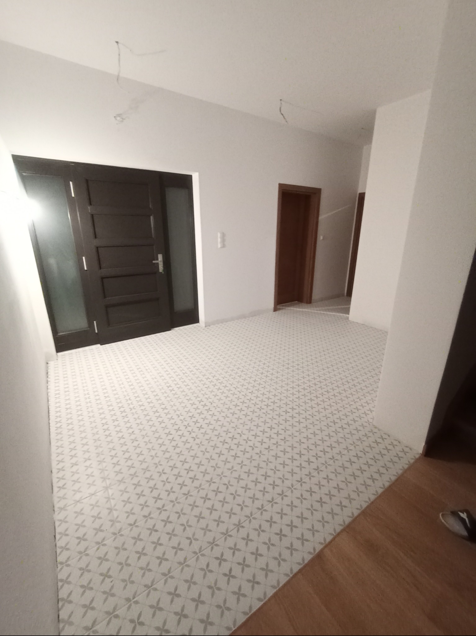 Wnętrze nowo wykończonego przedpokoju z białą ścianą, ciemnymi drzwiami wejściowymi i wzorzystą podłogą w odcieniach szarości, widoczne niedokończone przewody elektryczne na suficie.