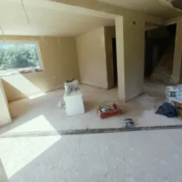 Wnętrze w trakcie tynkowania maszynowego, widoczne ściany pokryte świeżym tynkiem, narzędzia w czerwonym pojemniku, worki z zaprawą i niedokończone schody w tle.