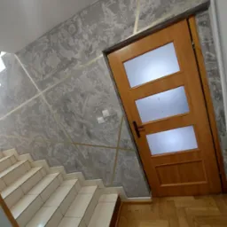 Ściana z dekoracyjnym tynkiem w odcieniach szarości, zdobiona złotymi paskami, sąsiadująca z drewnianymi schodami i drzwiami z trzema przeszkleniami.