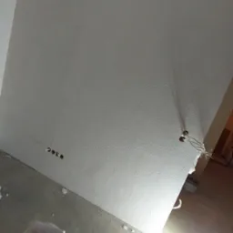 Ściana w trakcie prac wykończeniowych, częściowo pokryta gładzią, widoczny wałek malarski w wiadrze i lampa budowlana oświetlająca obszar roboczy.