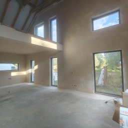 Usługi Ogólnobudowlane GRODEK - Surowe wnętrze domu z wysokim stropem i dużymi oknami po nałożeniu tynku. Widoczne instalacje pod sufitem i materiały budowlane na podłodze. Ściany w jasnym, szarym kolorze.