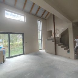 Usługi Ogólnobudowlane GRODEK - Surowe wnętrze domu w trakcie wykończenia: ściany po tynkowaniu, betonowa posadzka, schody w stanie surowym, duże okna z widokiem na zieleń. Widoczny pracownik w tle.