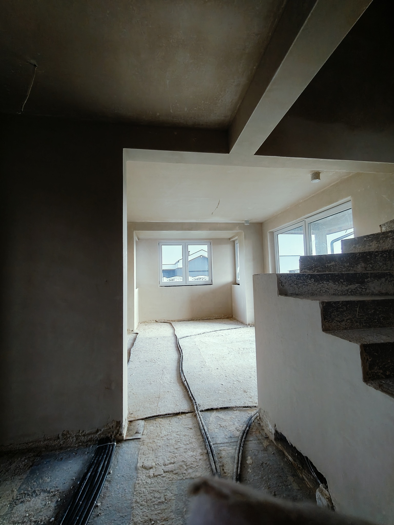 Surowe wnętrze w trakcie prac wykończeniowych: widoczne ściany po nałożeniu tynku, podłoga zabezpieczona folią i przewodami, schody w stanie surowym, okna z widokiem na budynki.