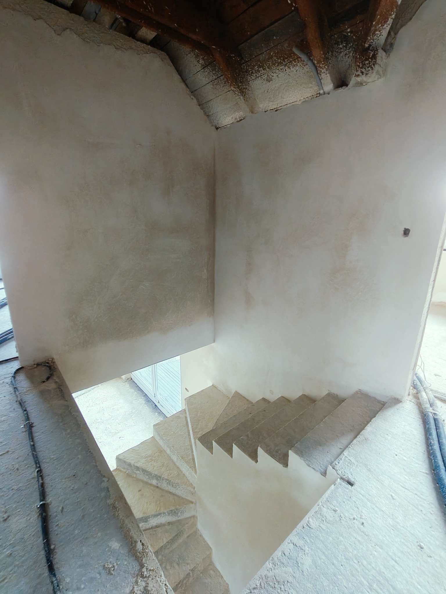 Wnętrze w trakcie remontu: świeżo otynkowane ściany klatki schodowej ze spiralnymi schodami, widoczne drewniane belki stropu i przewody instalacyjne.