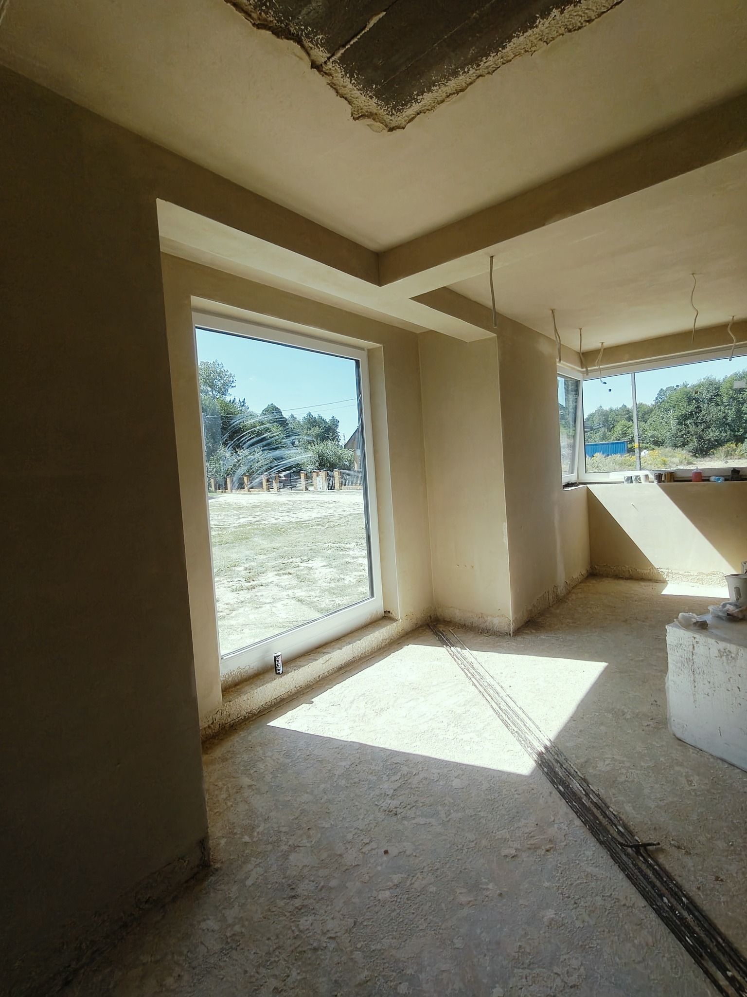 Wnętrze w stanie deweloperskim po nałożeniu tynków, widoczne okno z zewnątrz, surowy betonowy sufit z fragmentami starego tynku, przewody elektryczne zwisające z sufitu, podłoga z wylewką...
