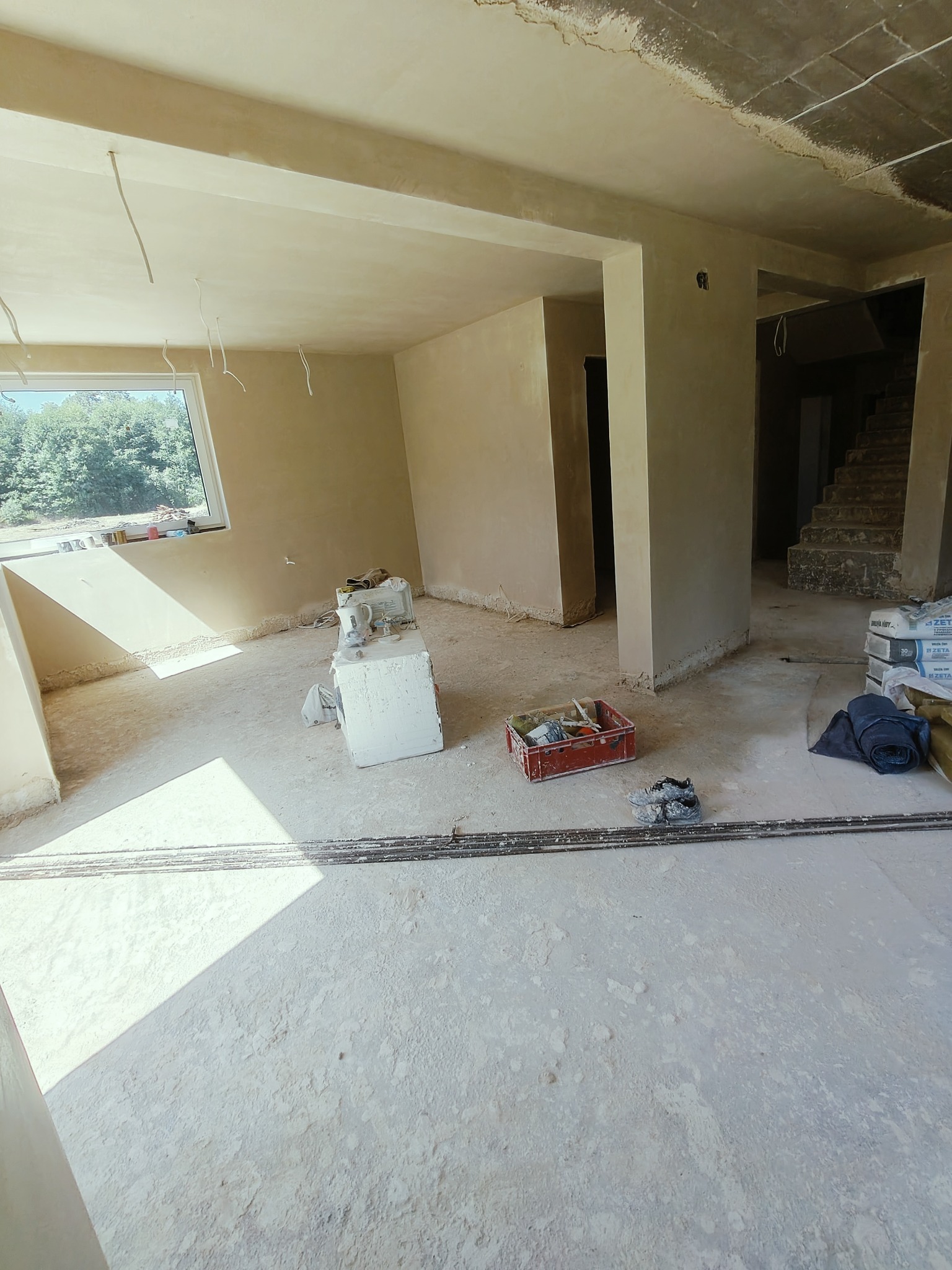 Wnętrze w trakcie tynkowania maszynowego, widoczne ściany pokryte świeżym tynkiem, narzędzia w czerwonym pojemniku, worki z zaprawą i niedokończone schody w tle.