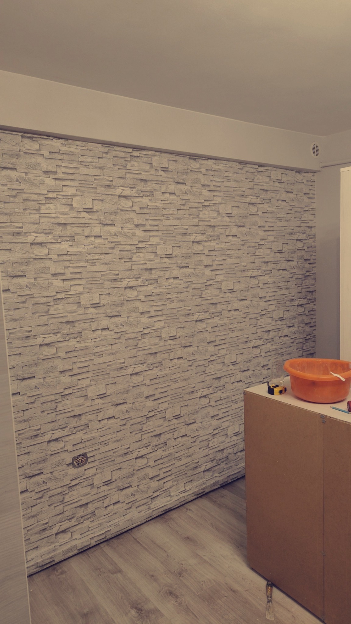 Ściana wykończona panelami imitującymi kamień, widoczna puszka elektryczna, na podłodze panele, obok stoi wiadro i narzędzia.