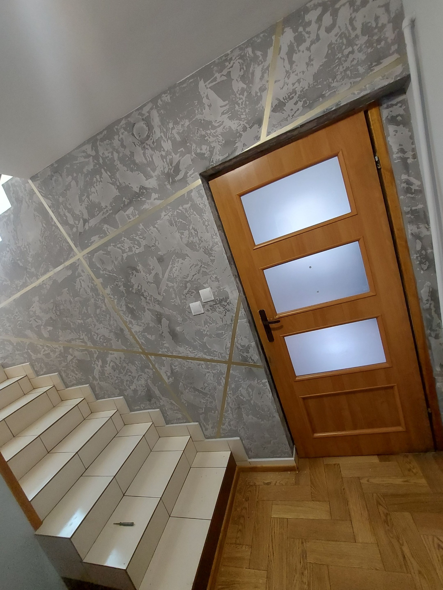 Ściana z dekoracyjnym tynkiem w odcieniach szarości, zdobiona złotymi paskami, sąsiadująca z drewnianymi schodami i drzwiami z trzema przeszkleniami.