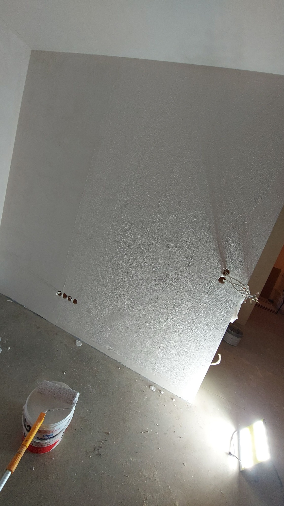 Ściana w trakcie prac wykończeniowych, częściowo pokryta gładzią, widoczny wałek malarski w wiadrze i lampa budowlana oświetlająca obszar roboczy.