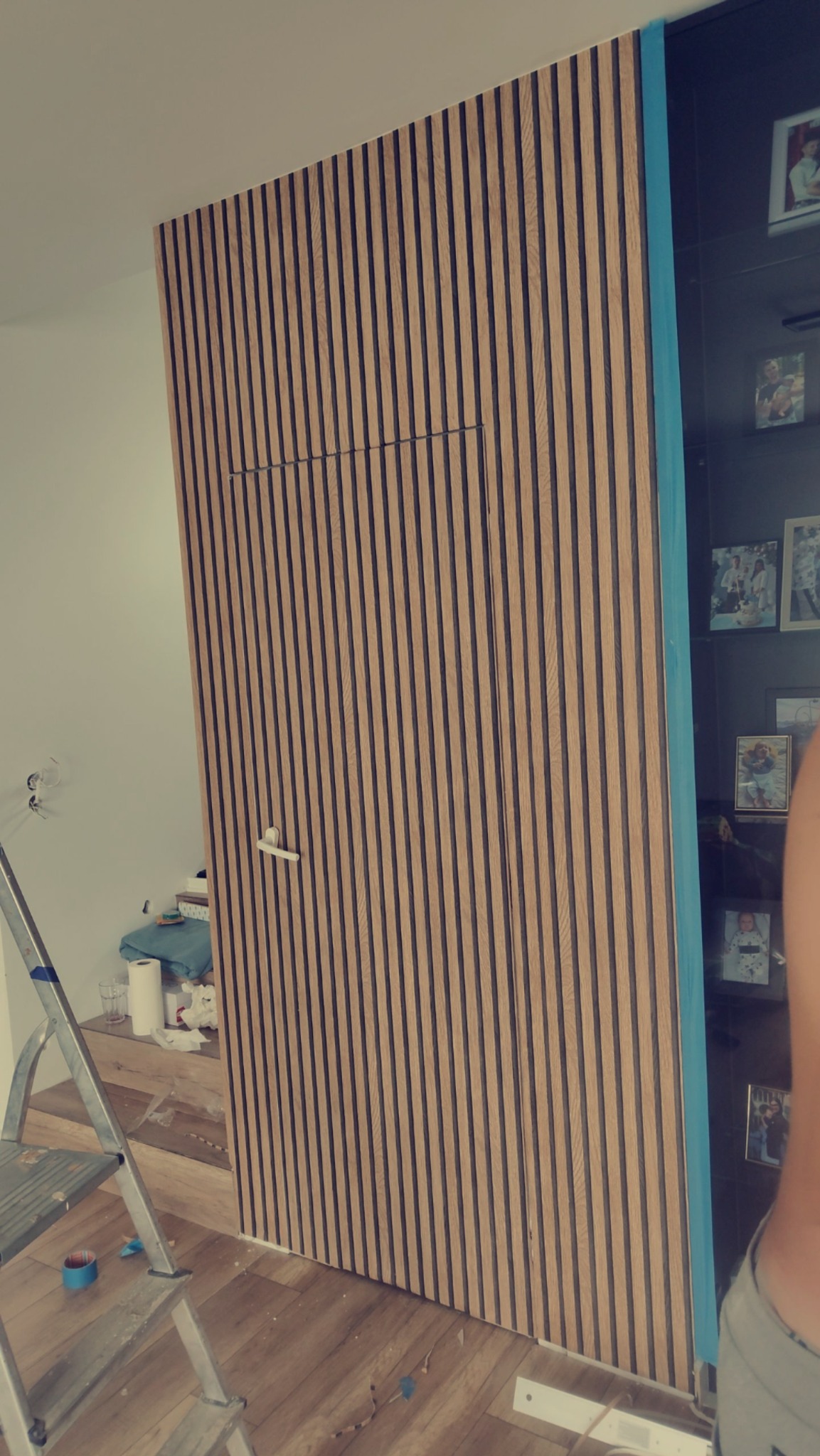 Drewniane lamele ścienne z ukrytymi drzwiczkami rewizyjnymi i niebieskim zabezpieczeniem przy szklanej witrynie, widoczna drabina i elementy montażowe na podłodze.