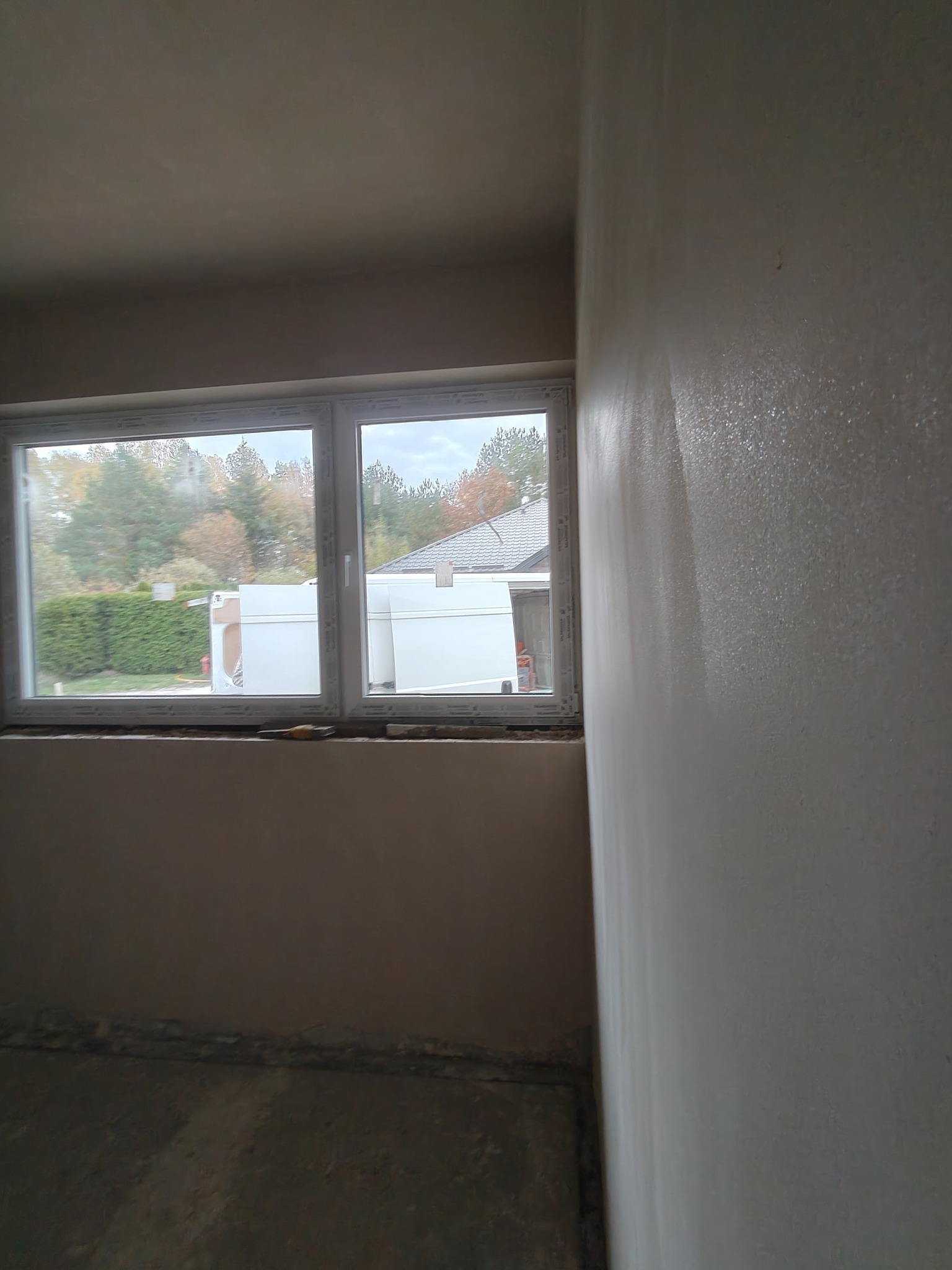 Świeżo nałożona, mokra gładź na ścianie w pokoju z oknem. Widoczne narzędzia pod oknem. Za oknem zieleń i budynek. Ściana w trakcie schnięcia.