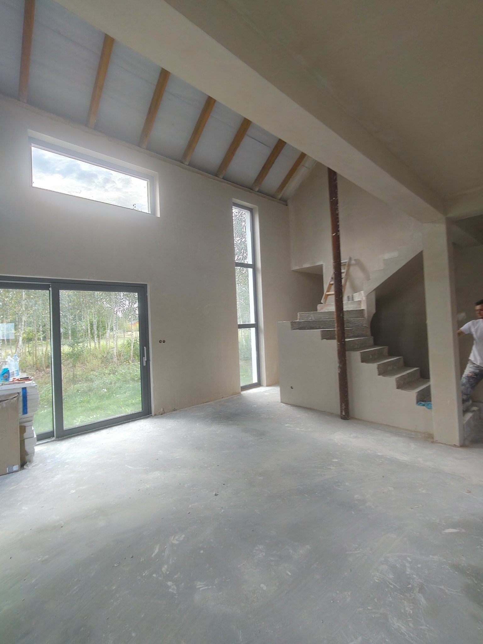 Surowe wnętrze domu w trakcie wykończenia: ściany po tynkowaniu, betonowa posadzka, schody w stanie surowym, duże okna z widokiem na zieleń. Widoczny pracownik w tle.