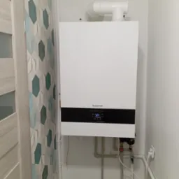 Biały piec gazowy marki Buderus zamontowany na ścianie, widoczne rury doprowadzające media, ściana z dekoracyjnymi panelami 3D w odcieniach bieli i turkusu po lewej stronie.