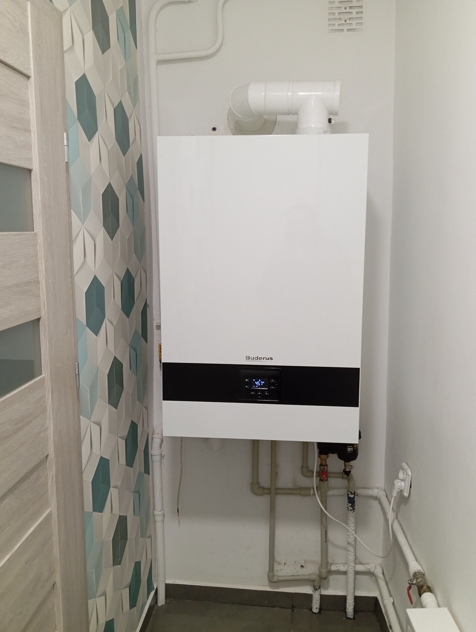 Biały piec gazowy marki Buderus zamontowany na ścianie, widoczne rury doprowadzające media, ściana z dekoracyjnymi panelami 3D w odcieniach bieli i turkusu po lewej stronie.