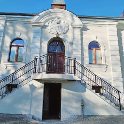 Firma Budowlana ROBI - Odnowiona elewacja budynku z białymi ścianami, okrągłymi oknami i schodami prowadzącymi do wejścia głównego nad piwnicą, widoczna tablica z napisem 'Sprottau 1908'.