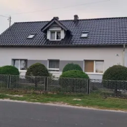 Dom jednorodzinny z ciemną dachówką i oknami dachowymi, otynkowany na szaro, z metalowym ogrodzeniem i kulistymi krzewami wzdłuż frontu.