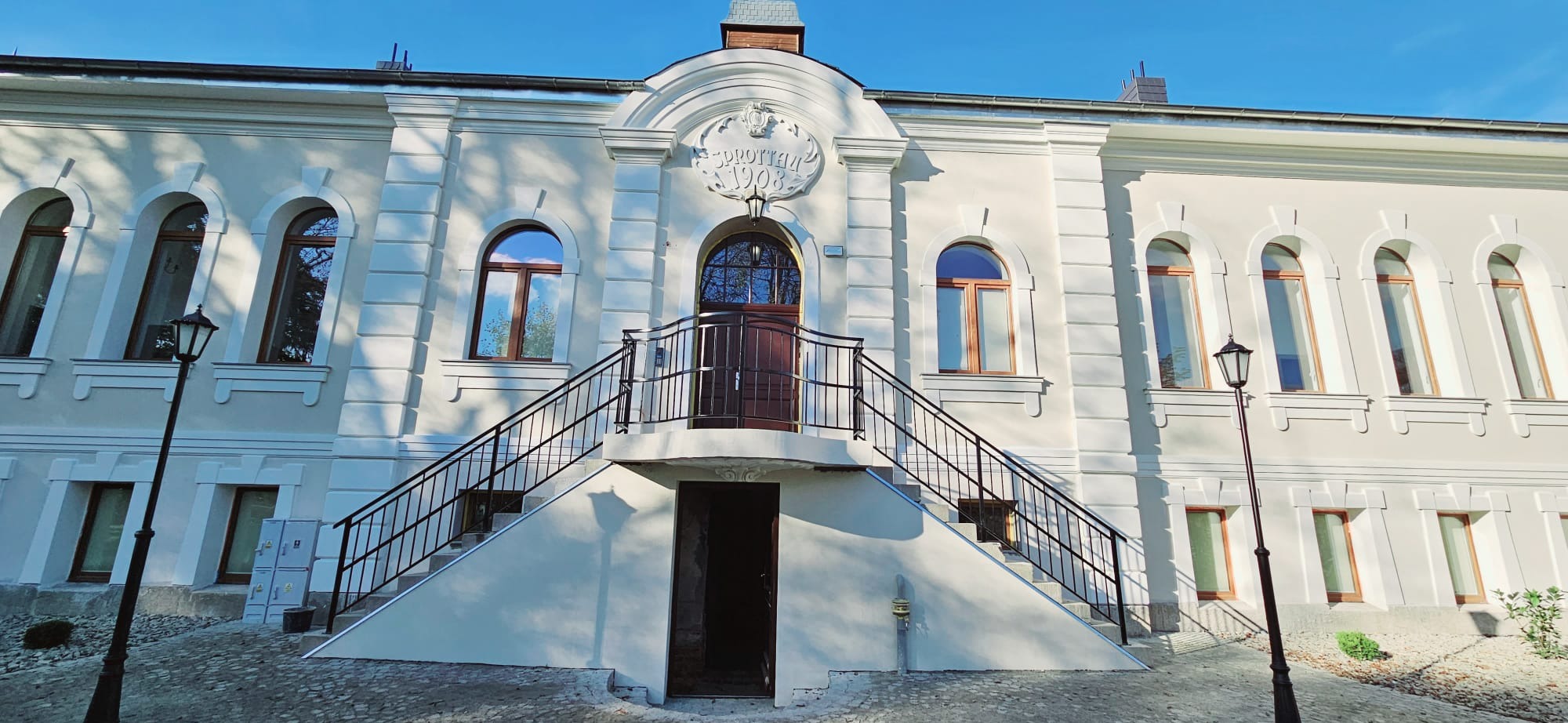 Odnowiona elewacja budynku z białymi ścianami, okrągłymi oknami i schodami prowadzącymi do wejścia głównego nad piwnicą, widoczna tablica z napisem 'Sprottau 1908'.