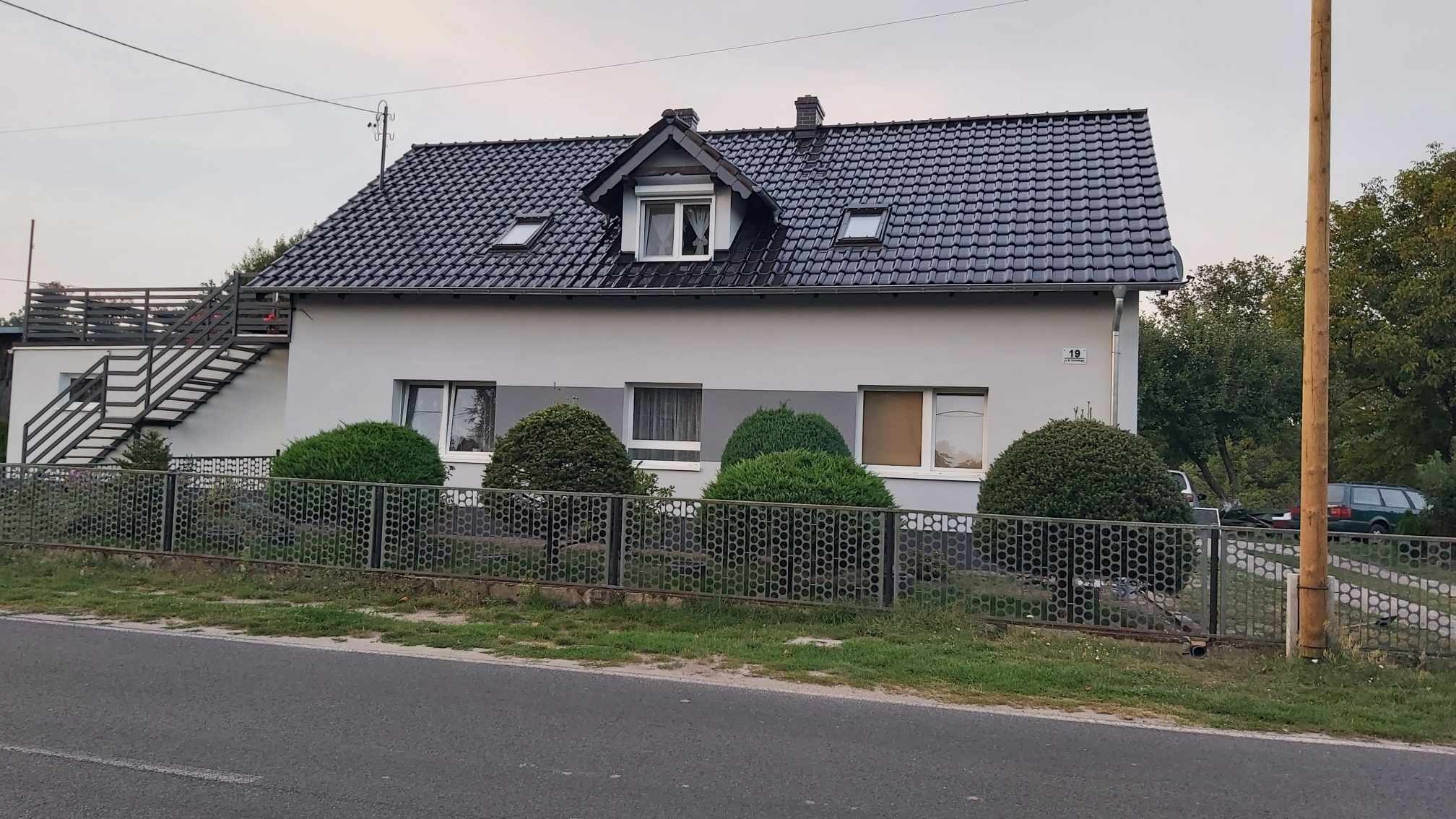 Dom jednorodzinny z ciemną dachówką i oknami dachowymi, otynkowany na szaro, z metalowym ogrodzeniem i kulistymi krzewami wzdłuż frontu.