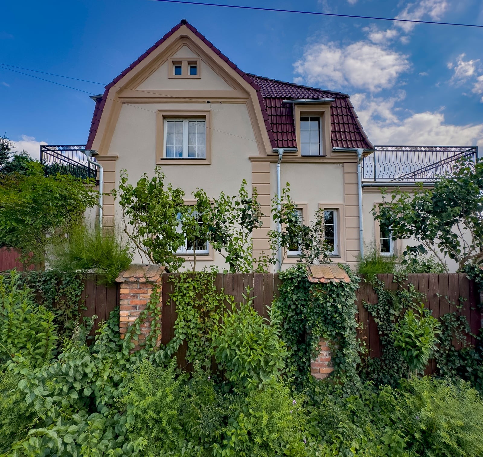 Elewacja domu z beżową fasadą, czerwonym dachem i balkonem, częściowo zasłonięta zielenią z drewnianym ogrodzeniem w tle.