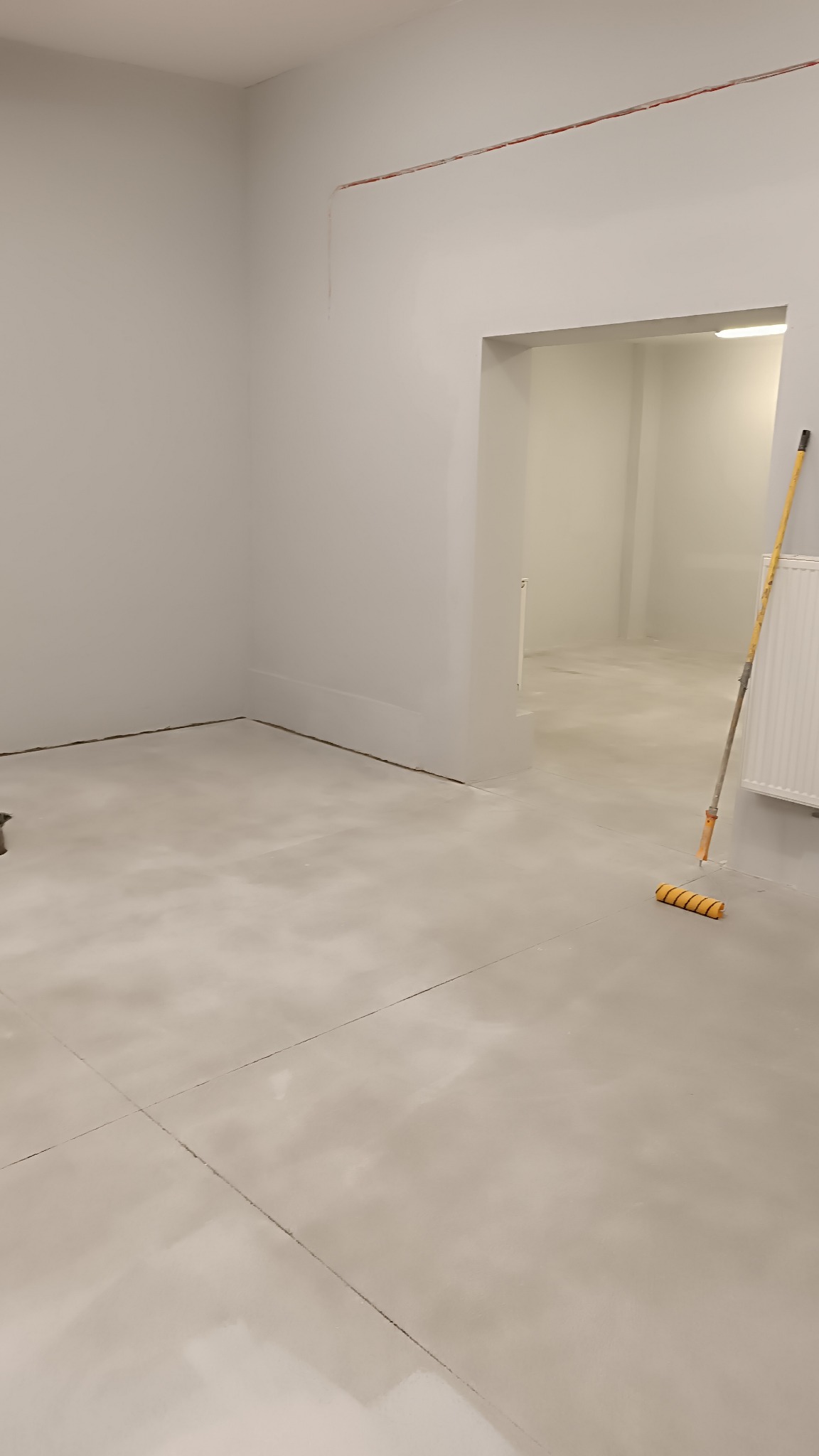 Świeżo pomalowane na szaro pomieszczenie z widocznym wałkiem do malowania opartym o grzejnik, na ścianie widoczne wystające przewody.