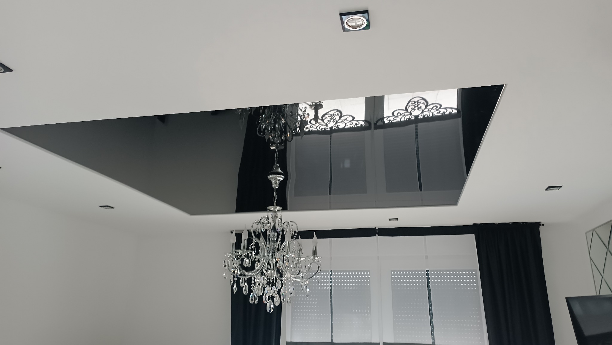 Nowoczesny salon z czarnym, lustrzanym sufitem podwieszanym i kryształowym żyrandolem. Widoczne punktowe oświetlenie i okna z czarnymi zasłonami.
