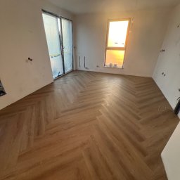 Onika Sp. z o.o. - Jasne panele ułożone w jodełkę w pustym, jasnym pokoju z oknem, częściowo wykończone ściany, przygotowane do dalszych prac.