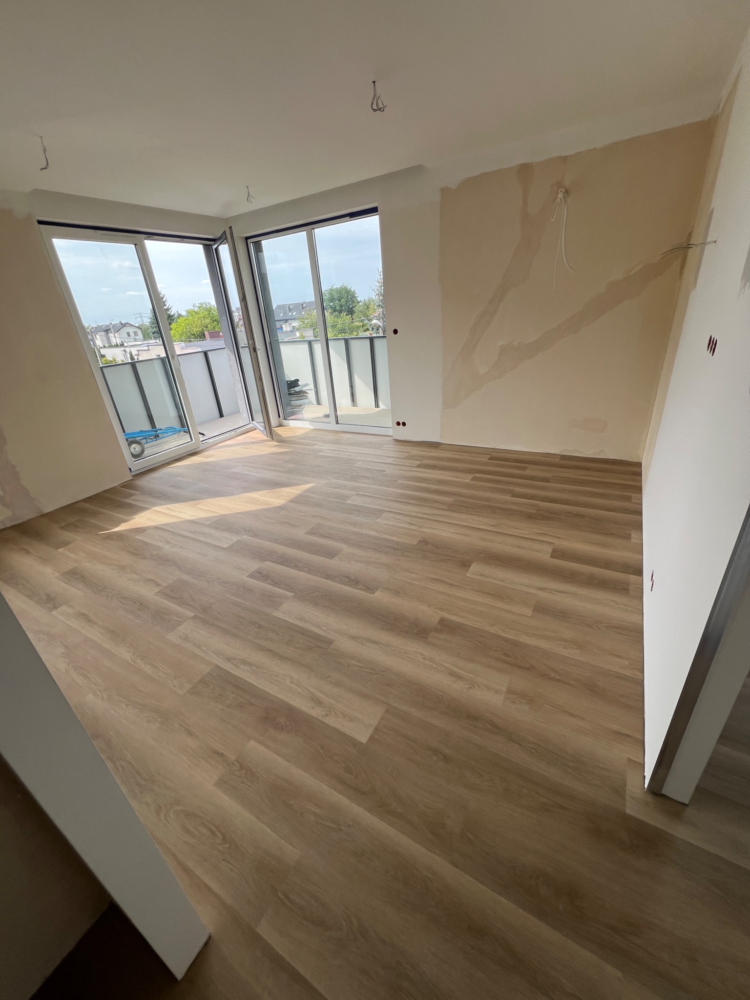Jasne panele podłogowe ułożone w nowym pokoju z dużym oknem balkonowym; ściany w trakcie wykańczania, widoczne przewody elektryczne.
