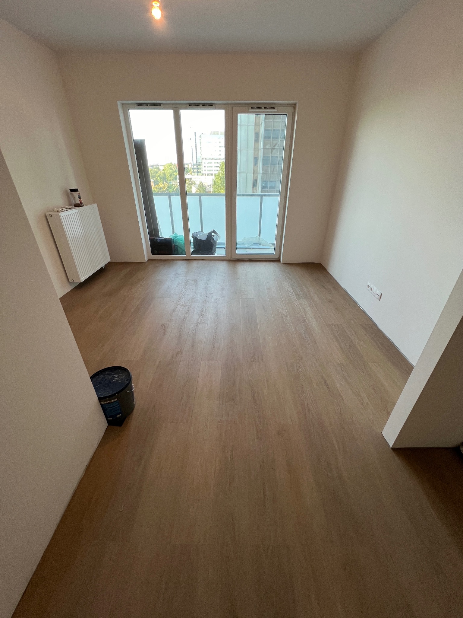 Pokój po położeniu paneli podłogowych, widoczny kubeł z farbą przy ścianie i grzejnik. Za oknem widok na miasto.