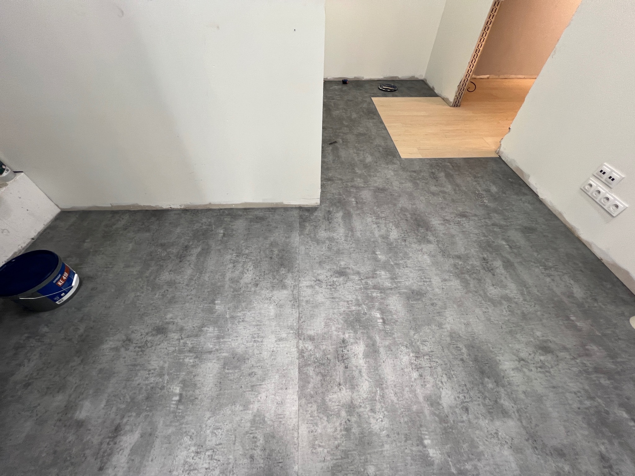 Szara wykładzina podłogowa o teksturze betonu w trakcie montażu, widoczny fragment surowej podłogi w przejściu i niebieskie wiadro z klejem przy ścianie.