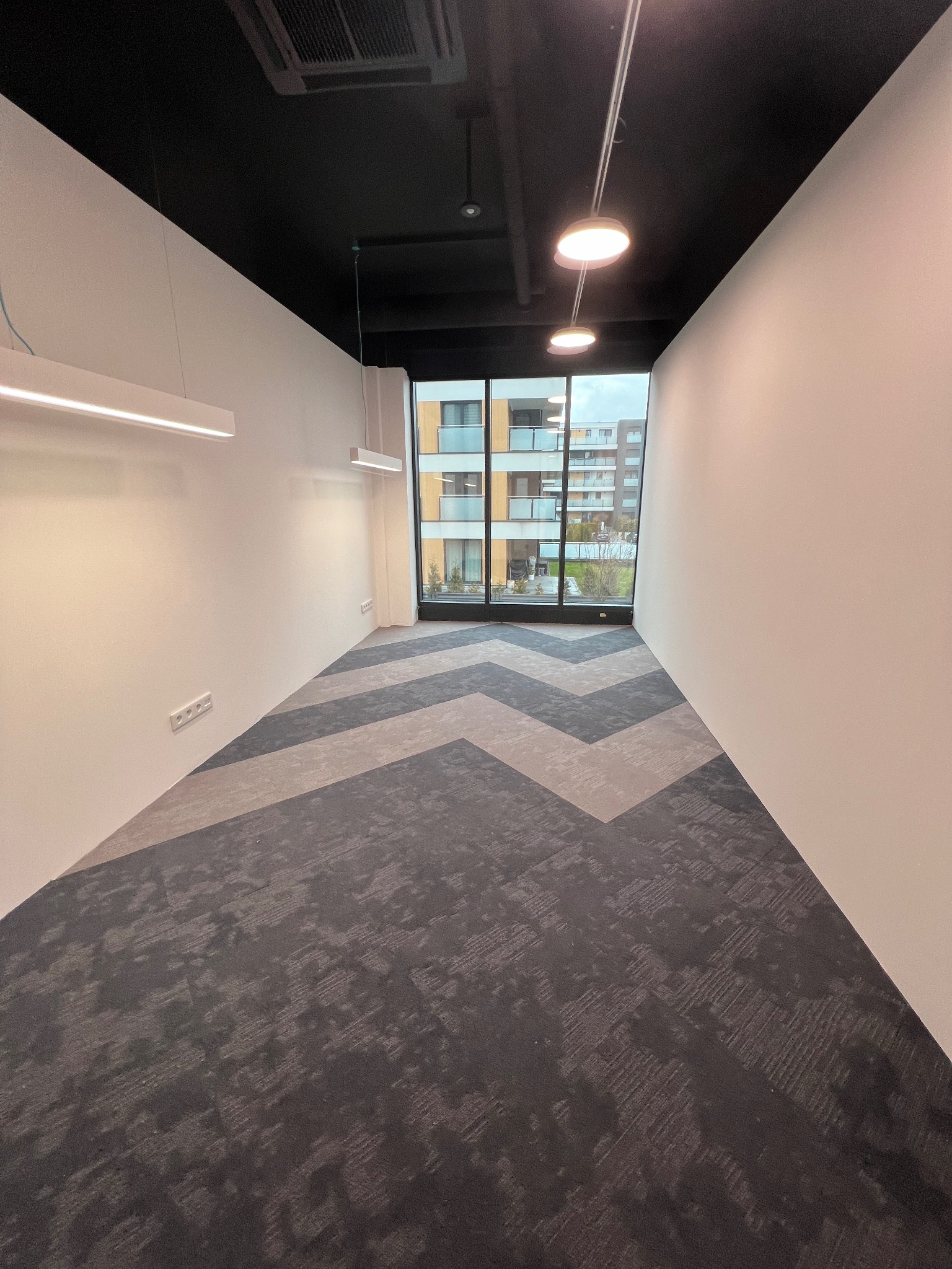 Pusta przestrzeń biurowa z nową wykładziną w zygzaki, widok na budynek mieszkalny przez okno, białe ściany i czarny sufit z oświetleniem LED.