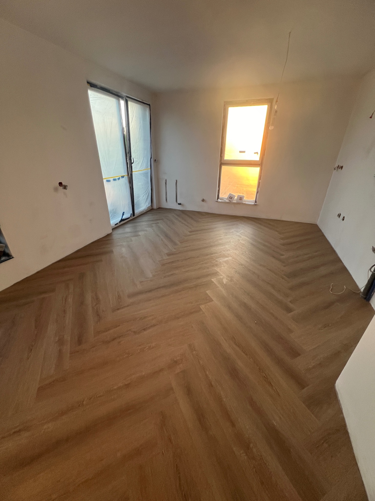 Jasne panele ułożone w jodełkę w pustym, jasnym pokoju z oknem, częściowo wykończone ściany, przygotowane do dalszych prac.