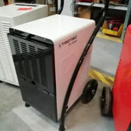 Biały osuszacz powietrza Trotec TTK 170 ECO z czarnymi elementami i uchwytem, stojący na kółkach w pomieszczeniu magazynowym, obok widoczny inny osuszacz w kolorze czerwonym.