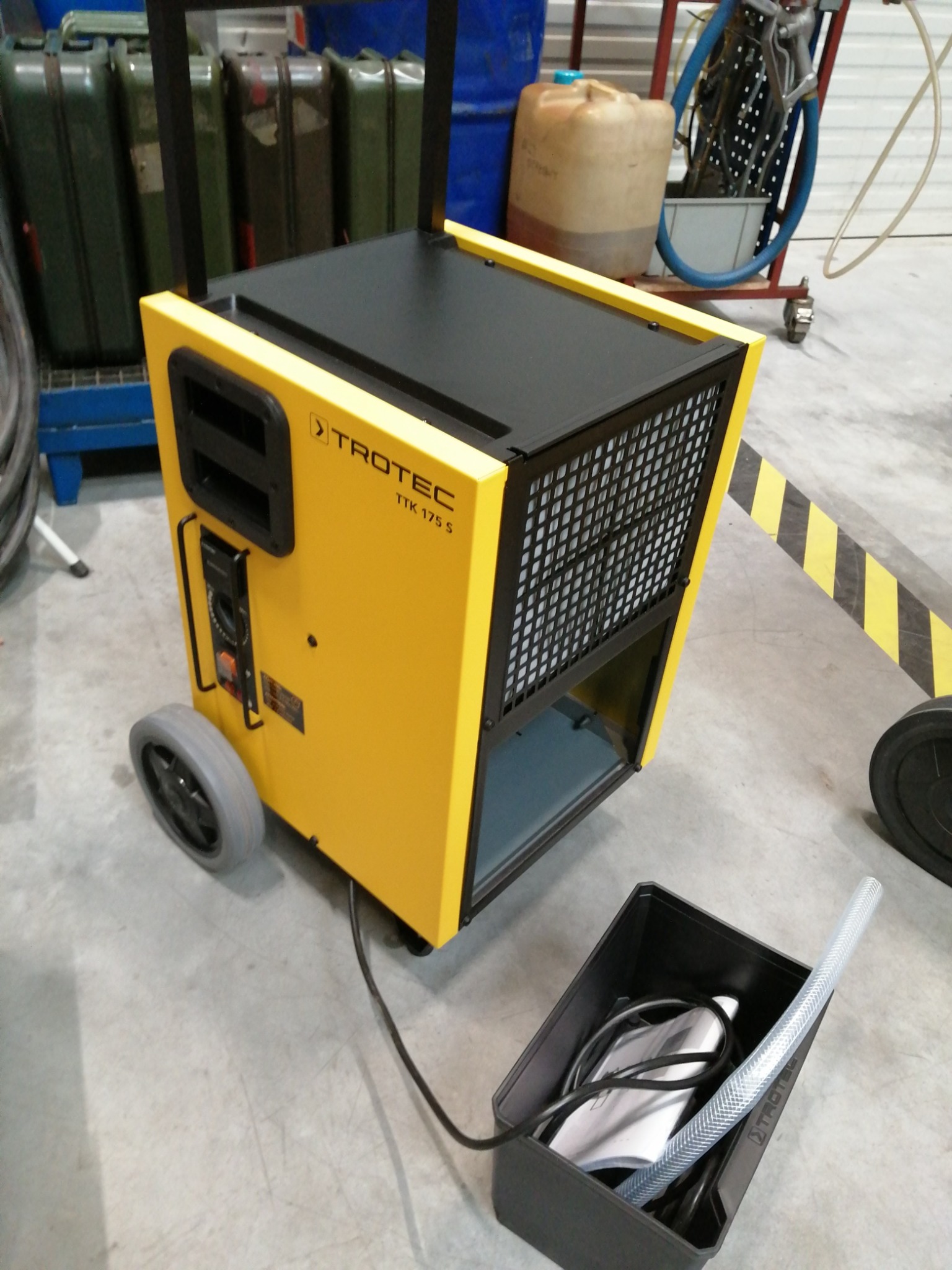 Żółty, mobilny osuszacz powietrza Trotec TTK 175 S z czarną kratką wentylacyjną i schowanymi akcesoriami w czarnym pojemniku, stojący na betonowej podłodze, w tle widoczne kanistry i inne urządzenia.