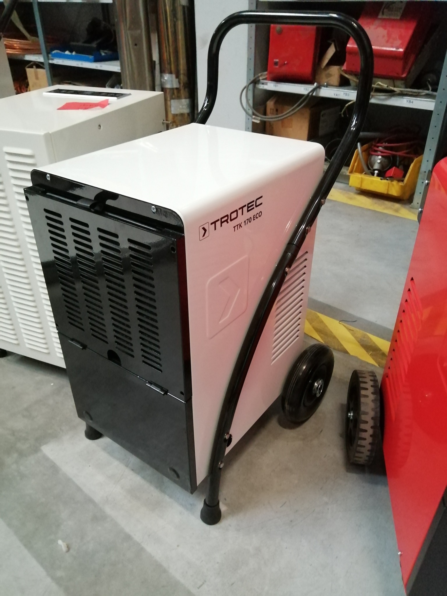 Biały osuszacz powietrza Trotec TTK 170 ECO z czarnymi elementami i uchwytem, stojący na kółkach w pomieszczeniu magazynowym, obok widoczny inny osuszacz w kolorze czerwonym.