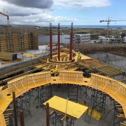 FUTURO - Widok z góry na plac budowy z żółtym szalunkiem, zbrojeniami i pracownikami w odblaskowych kamizelkach, w tle panorama miasta i morze pod pochmurnym niebem.
