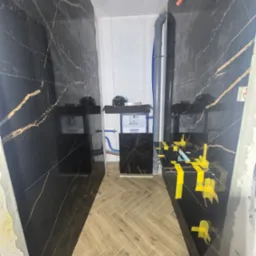 Wykończona łazienka w Paprotni z czarnymi płytkami imitującymi marmur ze złotymi żyłami, drewnopodobna podłoga ułożona w jodełkę, elementy instalacji widoczne.