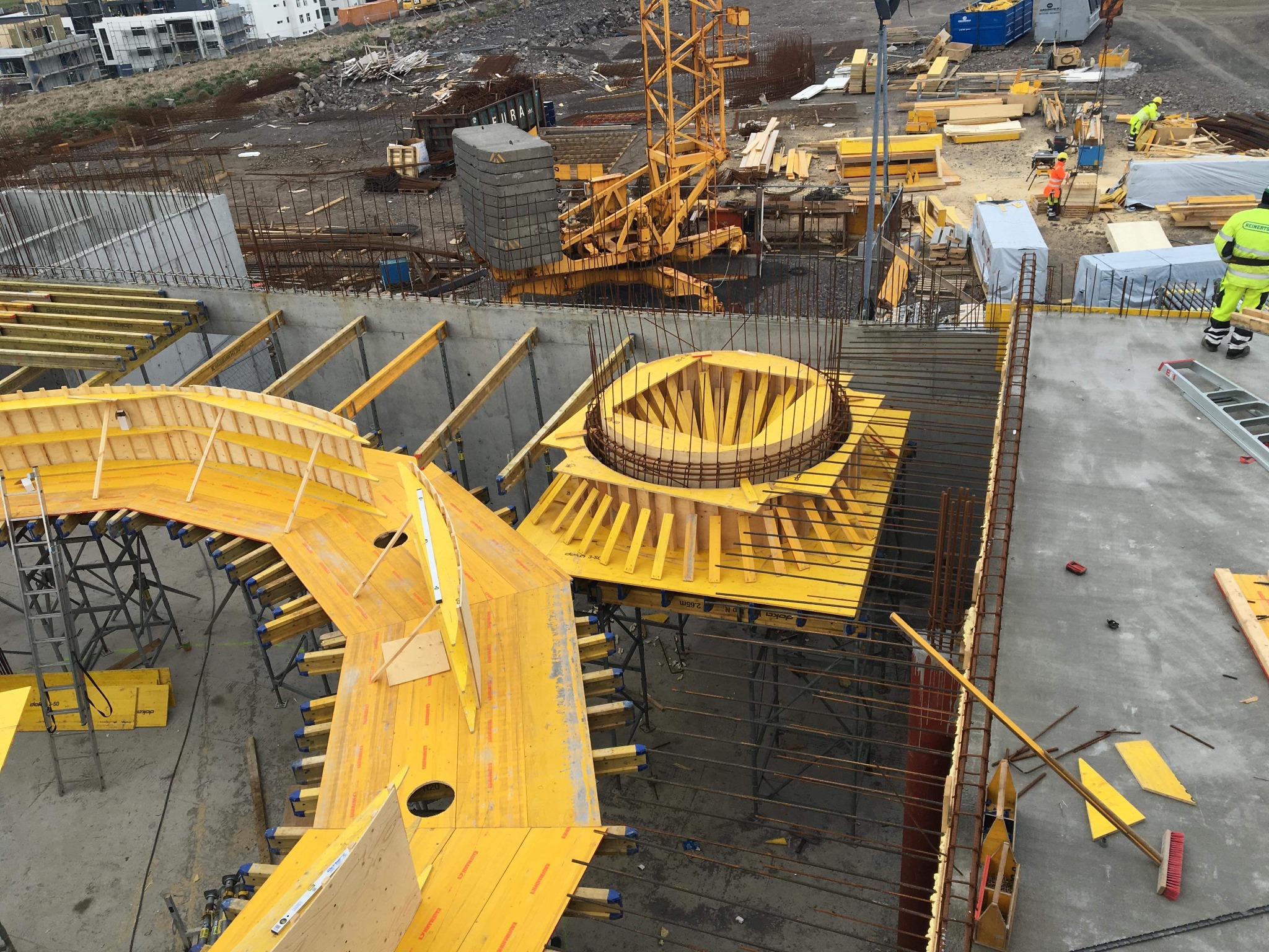 Widok z góry na plac budowy z żółtym szalunkiem do betonowania fundamentów o nietypowym kształcie, zbrojeniami i żurawiem w tle.