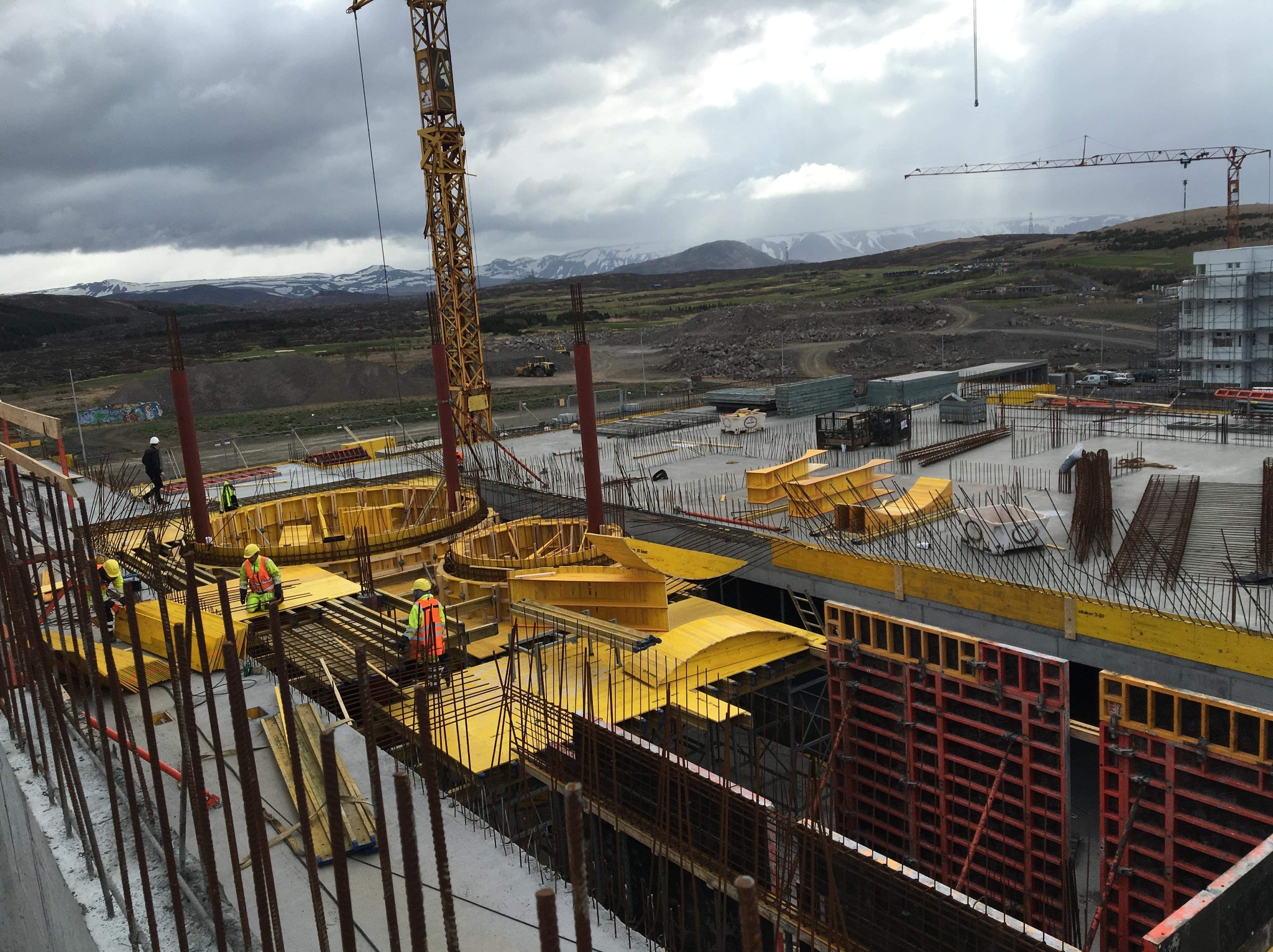 Budowa domu wielorodzinnego w trakcie, z widocznymi pracownikami w kaskach i kamizelkach odblaskowych układającymi szalunki i zbrojenie na tle krajobrazu z górami.