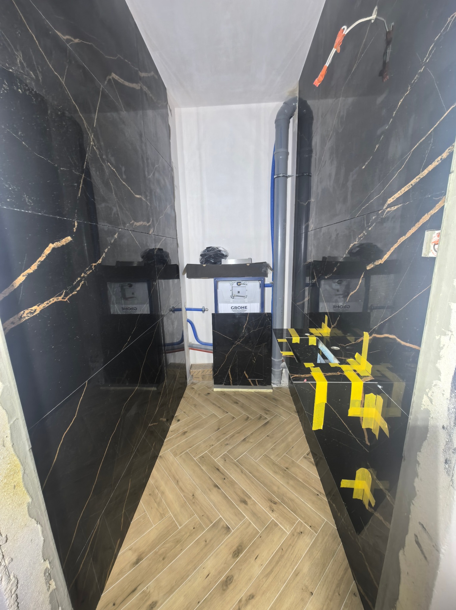 Wykończona łazienka w Paprotni z czarnymi płytkami imitującymi marmur ze złotymi żyłami, drewnopodobna podłoga ułożona w jodełkę, elementy instalacji widoczne.