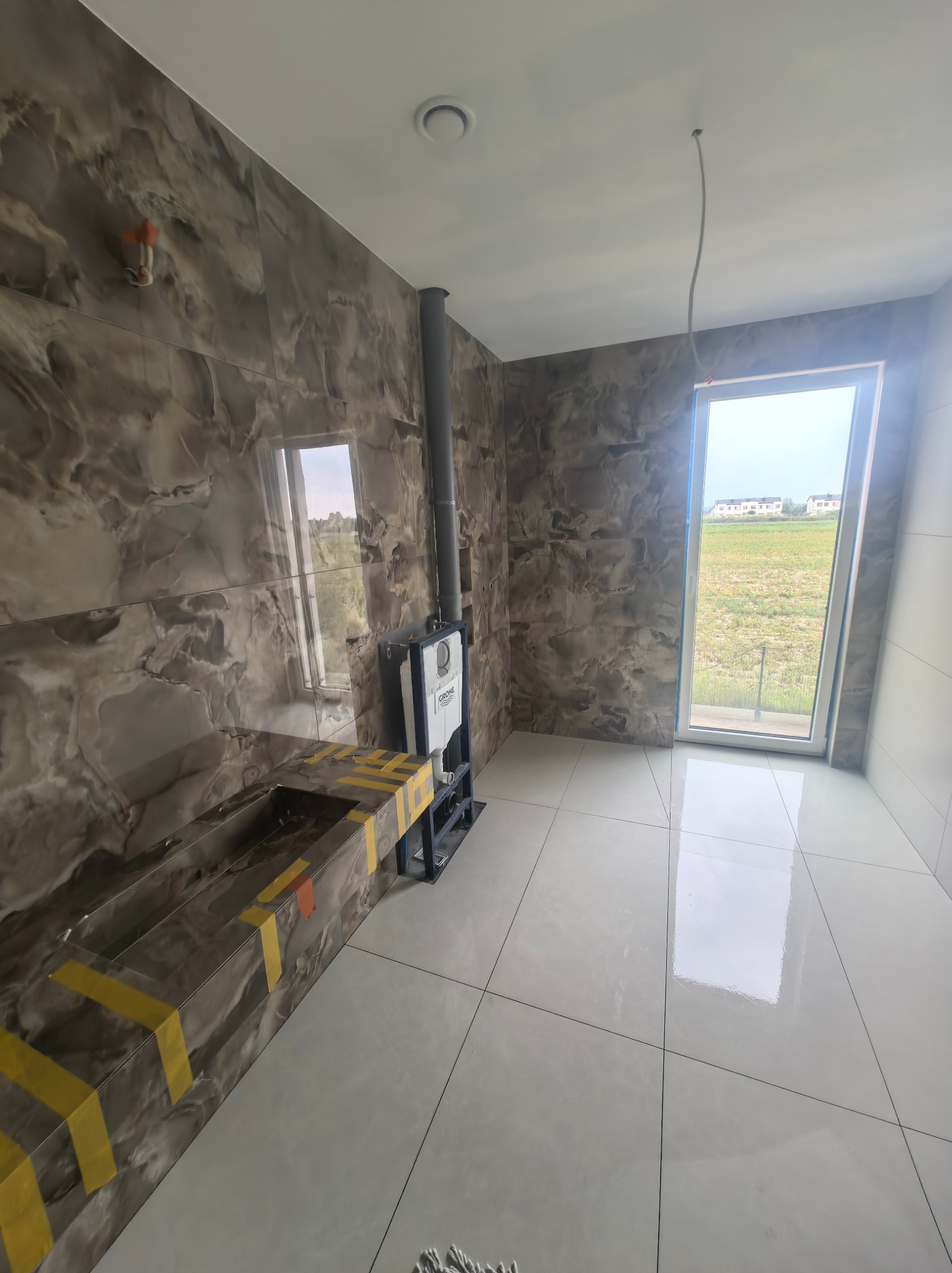 Wykończona łazienka z marmurowymi płytkami, zabudową pod umywalkę z żółtą taśmą zabezpieczającą i widokiem na pole przez okno.