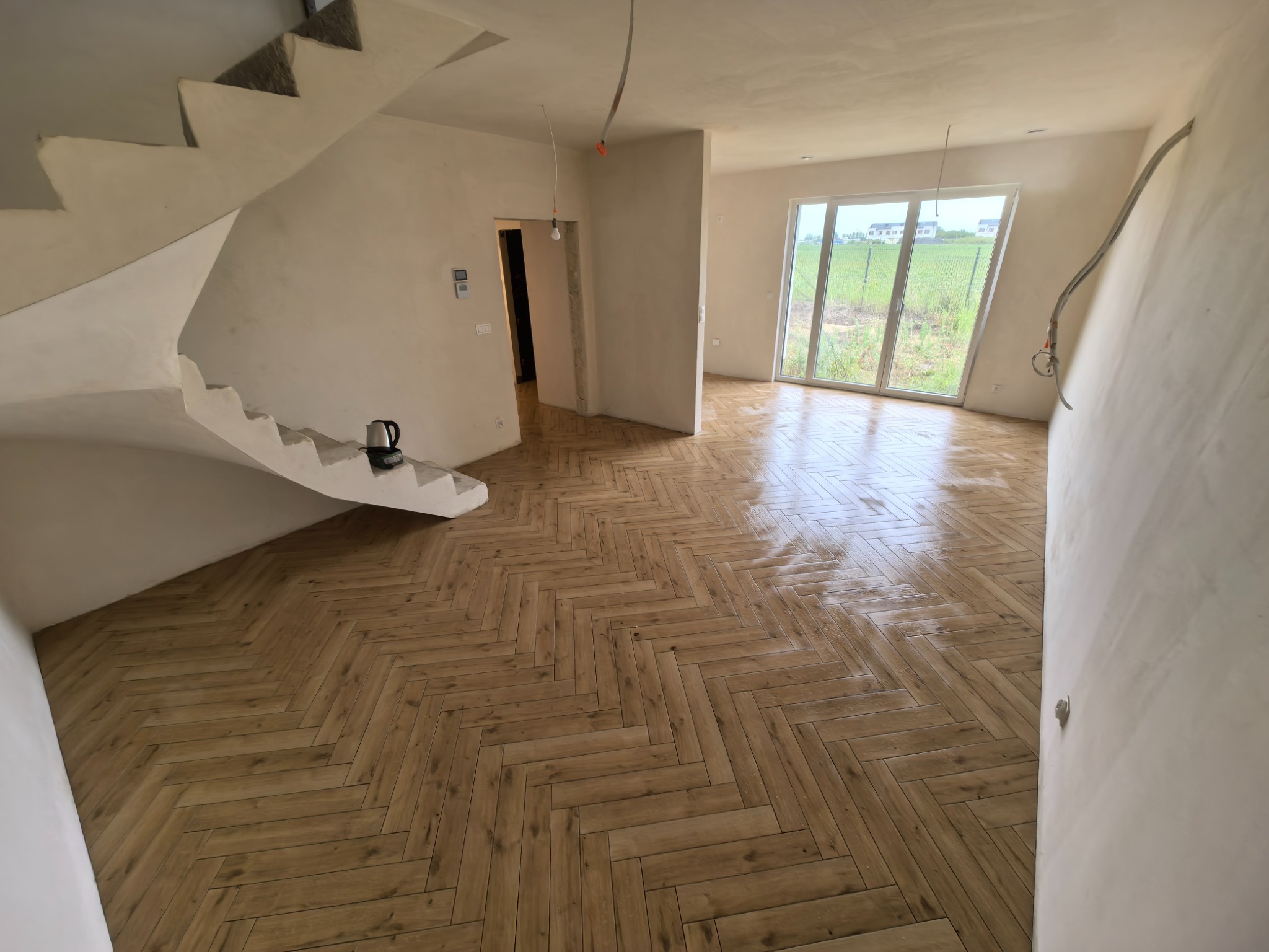 Jasne, przestronne wnętrze z ułożoną parkietową podłogą w jodełkę, spiralnymi schodami i dużym oknem z widokiem na zieleń. Ściany w surowym stanie, widoczne instalacje.