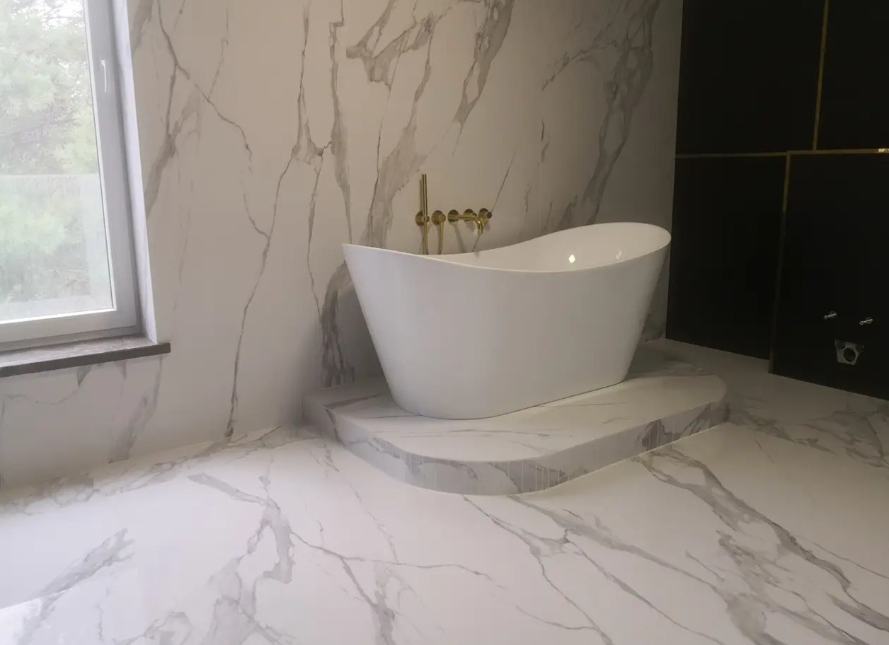 Nowoczesna łazienka z wolnostojącą wanną na podeście, wykończona płytkami imitującymi marmur i czarnymi panelami. Złota armatura dodaje elegancji.