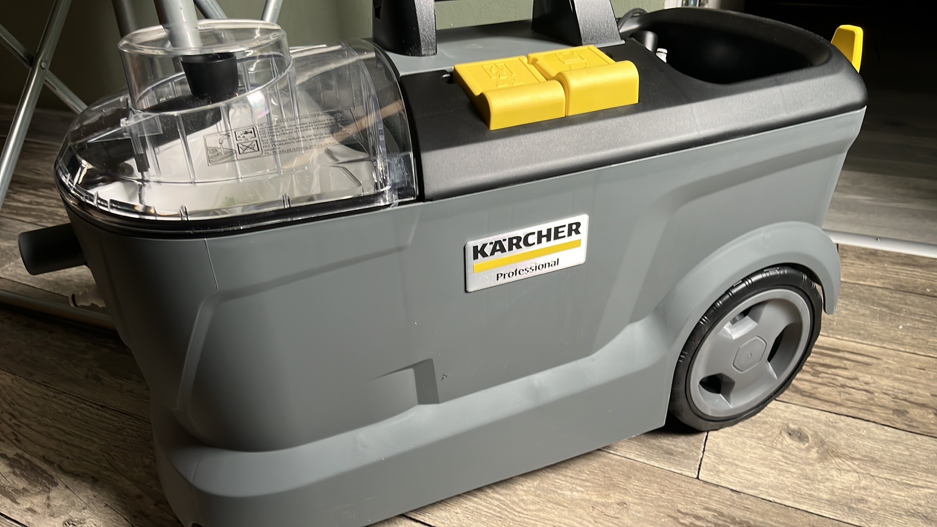 Szary odkurzacz piorący Kärcher Professional z przezroczystym zbiornikiem na wodę, widoczny na drewnianej podłodze, z żółtymi elementami mocującymi.